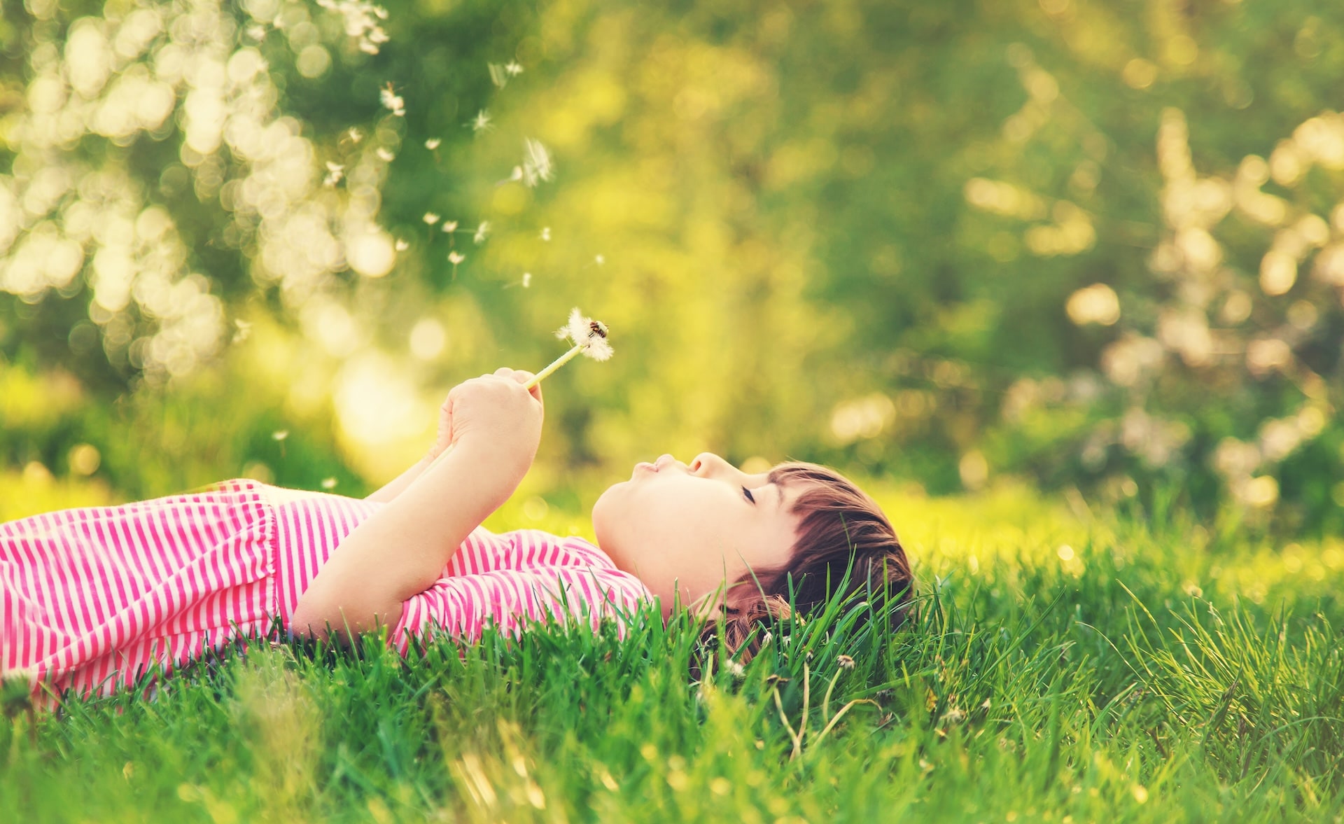 Ein Kind liegt entspannt im Gras und pustet eine Pusteblume – Symbol für die spielerische, sanfte Wirkung von Kinderhypnose